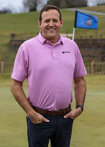 Bryan Kraushofer - Carroll News | Carroll Publications | Carroll County, MD Man in a pink polo shirt standing on a golf course green with a blue flag in the background, smiling with hands in pockets.