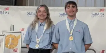 Two smiling SkillsUSA competitors wearing blue shirts with gold medals on stage beside a first-place sign.