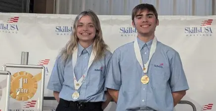 Two smiling SkillsUSA competitors wearing blue shirts with gold medals on stage beside a first-place sign.