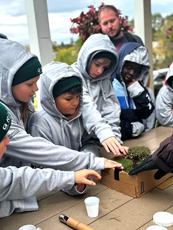 first-green-golf-children - Carroll News | Carroll Publications | Carroll County, MD Group of kids in gray hoodies gathered around a wooden box, planting or inspecting soil, under adult supervision outdoors.