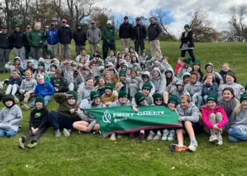 Large group of students and instructors posing on a grassy hill, holding a green First Green banner.