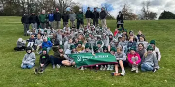 Large group of students and instructors posing on a grassy hill, holding a green First Green banner.
