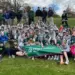 Large group of students and instructors posing on a grassy hill, holding a green First Green banner.