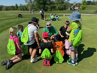 first-green-image-02 - Carroll News | Carroll Publications | Carroll County, MD Group of children sitting in a circle on a grassy field with bright green backpacks, listening to instructors at an outdoor camp.
