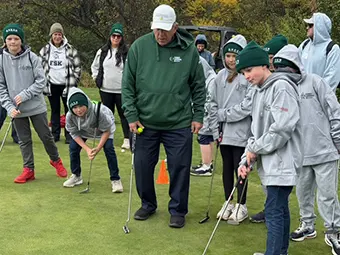 first-green-image - Carroll News | Carroll Publications | Carroll County, MD Youth golf lesson: coach demonstrates putting to a group on a practice green outside in fall colors organic setting.