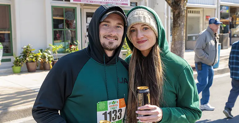 Two people in hoodies posing together at a street race, wearing bib 1133; the woman holds a can and smiles at the camera with sunny storefronts behind them in a small town.