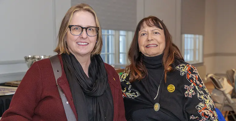 Two smiling women pose for a photo indoors, one wearing glasses and a burgundy cardigan, the other in a black top with a floral scarf.