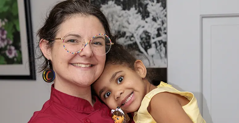 Smiling woman with colorful glasses hugs a young girl indoors, both looking at the camera.