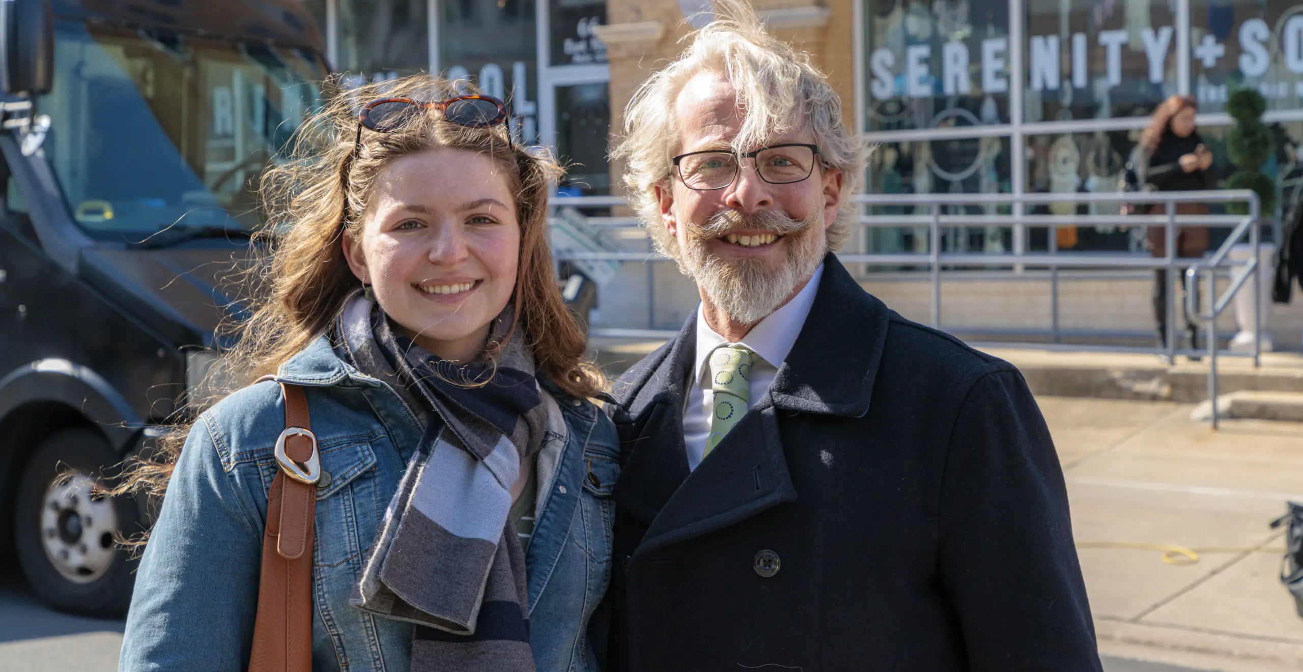 Young woman in a denim jacket and scarf standing beside an older man with glasses and a gray beard, both smiling in an urban plaza.