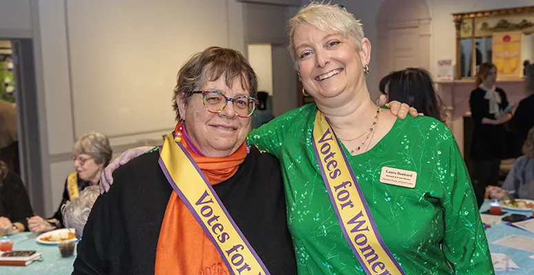 Two women at a community event pose together, wearing sashes that read 'Votes for Women' and smiling warmly.