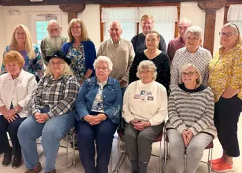 Group of fourteen seniors posing for a photo in a bright community room, with seven seated in front and seven standing behind, casual attire.