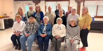 Group of fourteen seniors posing for a photo in a bright community room, with seven seated in front and seven standing behind, casual attire.
