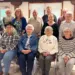 Group of fourteen seniors posing for a photo in a bright community room, with seven seated in front and seven standing behind, casual attire.
