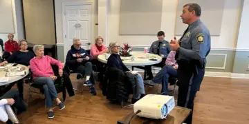 Senior adults seated at round tables in a community room, listening to a uniformed police officer giving a talk.