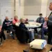 Senior adults seated at round tables in a community room, listening to a uniformed police officer giving a talk.
