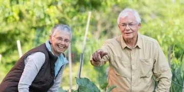Two smiling seniors gardening together in a lush outdoor yard.