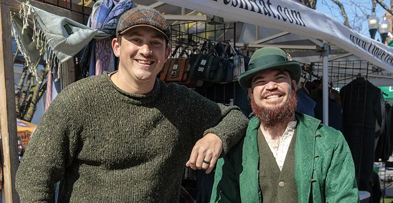 Two men stand at an outdoor market stall, one in a dark knit sweater and cap, the other in a green suit and hat, smiling at the camera amid hanging bags and clothing.