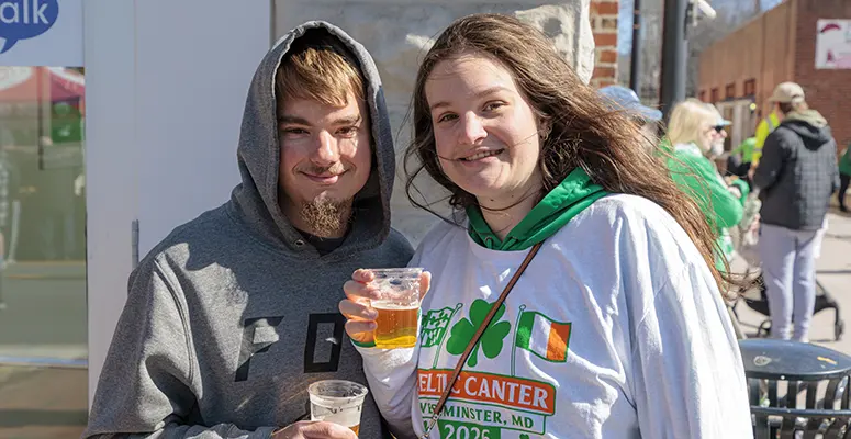 Two smiling people pose outdoors, the man in a gray hoodie and the woman in a white event shirt, each holding a cup of drinks for a fundraiser.