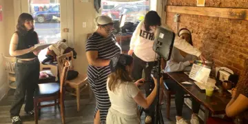 Group of people filming a video in a cafe, with a camera on a tripod and a monitor setup in the center of the scene.