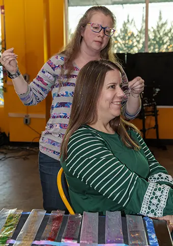 Hairdresser styles a client’s hair while the client smiles, seated at a table with colorful ribbons on display.