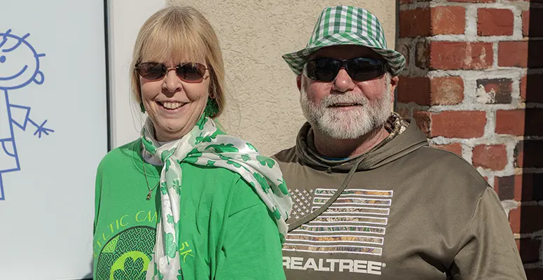 Two adults smile for the camera outdoors, wearing green clothing and sunglasses; a woman in a green shirt with a white scarf, a man in a brown tee and green bucket hat.