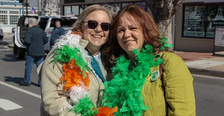 Two women smile for a photo at a street parade, wearing bright green and orange feather boas over their jackets.
