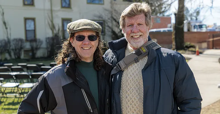 Two smiling adults standing outside on a sunny day, wearing jackets; man has a shoulder bag strap across his chest.