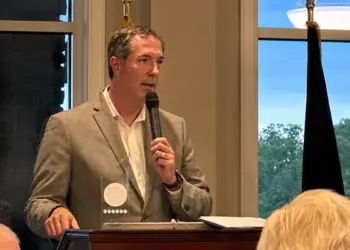 Man in a gray blazer speaks into a handheld microphone at a wooden podium during a formal event, with flags and attendees in the background.