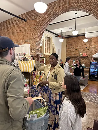 Woman in beige jacket speaks with a man pushing a shopping basket inside a brick-walled store.