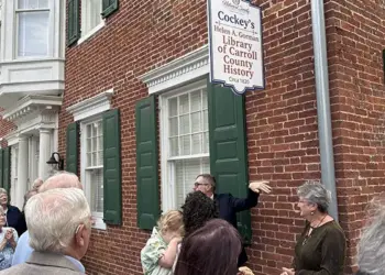 Crowd gathers outside a brick historic library building with green shutters and a hanging sign for Cockey's Helen B. Gorman Library of Carroll County History.