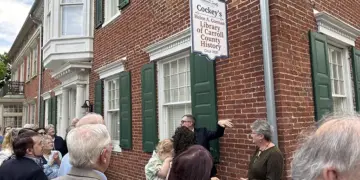 Crowd gathers outside a brick historic library building with green shutters and a hanging sign for Cockey's Helen B. Gorman Library of Carroll County History.