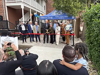 Group of people at a ribbon-cutting ceremony outside a building under a blue canopy, with reporters and smiling spectators taking photos.