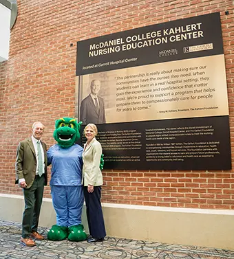 mcdaniel-college-foundation - Carroll News | Carroll Publications | Carroll County, MD Two adults in business attire stand with a green dragon mascot beside a large informational plaque mounted on a brick wall that reads McDaniel College Kahler Nursing Education Center and related text.