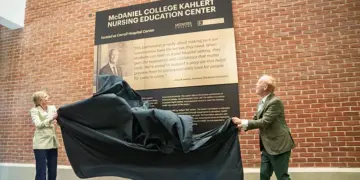 Two people unveil a plaque for the McDaniel College Kahler Nursing Education Center against a brick wall outside a hospital setting.