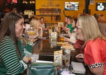 Two women sit across from each other at a long communal table in a lively restaurant, toasting with drinks and smiling.