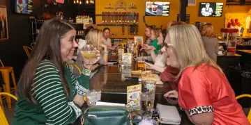 Two women sit across from each other at a long communal table in a lively restaurant, toasting with drinks and smiling.