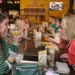 Two women sit across from each other at a long communal table in a lively restaurant, toasting with drinks and smiling.