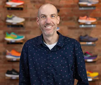 Smiling man in a navy patterned shirt stands in a sneaker shop with colorful shoes on brick wall shelves behind him.