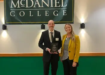 Two adults stand on a stage under a McDaniel College banner; man in a suit holds an award plaque, woman in a yellow blazer smiles beside him.