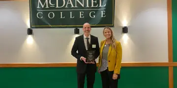 Two adults stand on a stage under a McDaniel College banner; man in a suit holds an award plaque, woman in a yellow blazer smiles beside him.