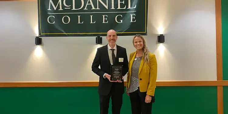 Two adults stand on a stage under a McDaniel College banner; man in a suit holds an award plaque, woman in a yellow blazer smiles beside him.