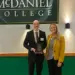 Two adults stand on a stage under a McDaniel College banner; man in a suit holds an award plaque, woman in a yellow blazer smiles beside him.