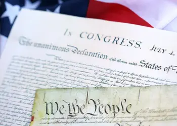 Close-up of the U.S. Declaration of Independence and a parchment reading 'We the People,' with an American flag in the background.