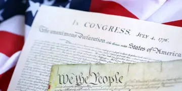 Close-up of the U.S. Declaration of Independence and a parchment reading 'We the People,' with an American flag in the background.
