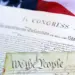 Close-up of the U.S. Declaration of Independence and a parchment reading 'We the People,' with an American flag in the background.