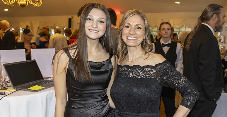 Two women in black evening dresses smile for a photo at a formal event, with guests and a laptop on a nearby table in the background.