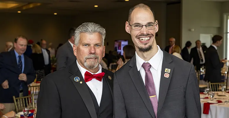 Two men in formal attire pose for a photo at a banquet, with guests and tables in the background.