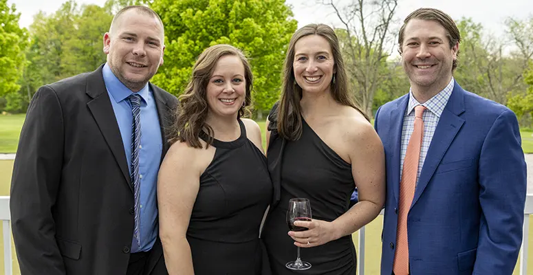 Four adults in formal attire posing together outdoors at a social event; two women in black dresses, two men in suits smiling with a wine glass visible.