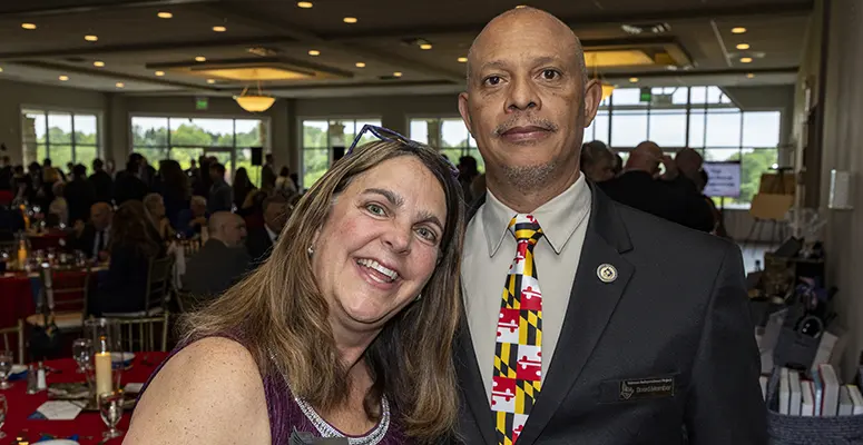 Smiling woman with glasses on her head posing with a man in a suit at a formal banquet, with tables and guests visible in the background.