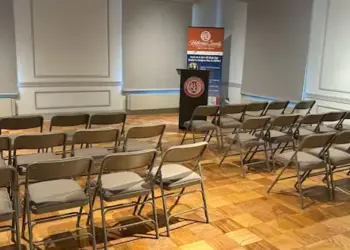 Empty conference room with rows of brown folding chairs facing a podium and banner at the front of the room.